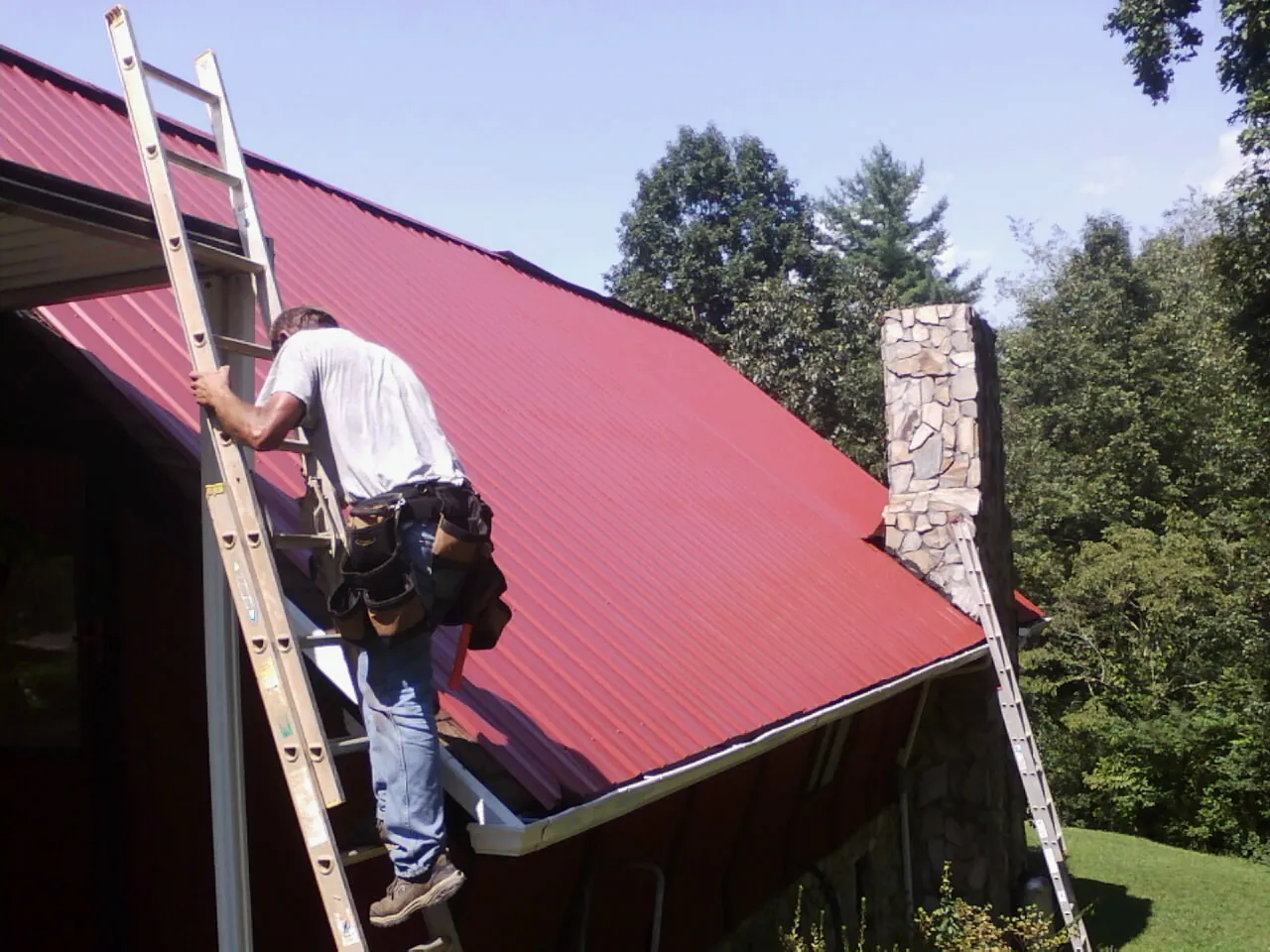 Roofer on a ladder beside a red metal roof and stone chimney — Soffit & Fascia Repair by Roofing Brothers in Burnside